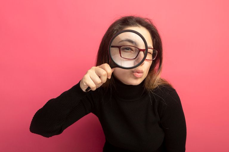 Young beautiful woman in a black turtleneck and glasses looking at front through magnifying glass standing over pink wall

