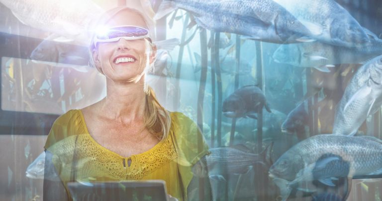 a woman in a yellow blouse with virtual reality goggles on. Virtual fish swimming around her.