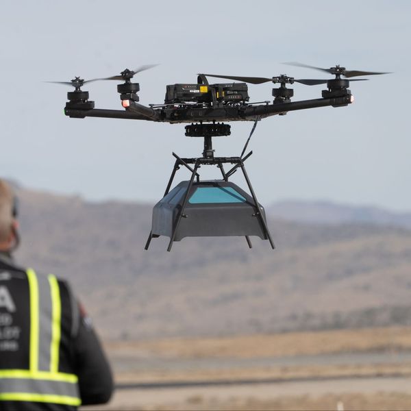 A drone in flight, carrying an antennae array payload beneath it. The drone has multiple rotors and is a large, professional-grade model designed for heavy lifting or specialized tasks. In the foreground, a person wearing a high-visibility vest with "FAA" and "Certified Remote Pilot" written on the back is controlling the drone. The background features an open, barren landscape with distant hills, suggesting a test or training area.