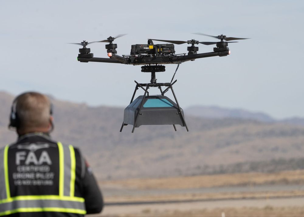 A drone in flight, carrying an antennae array payload beneath it. The drone has multiple rotors and is a large, professional-grade model designed for heavy lifting or specialized tasks. In the foreground, a person wearing a high-visibility vest with "FAA" and "Certified Remote Pilot" written on the back is controlling the drone. The background features an open, barren landscape with distant hills, suggesting a test or training area.