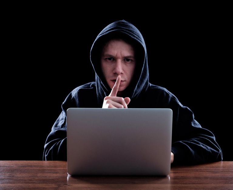 A man at his laptop in a hoodie sweater with his finger over his mouth making the shush motion.