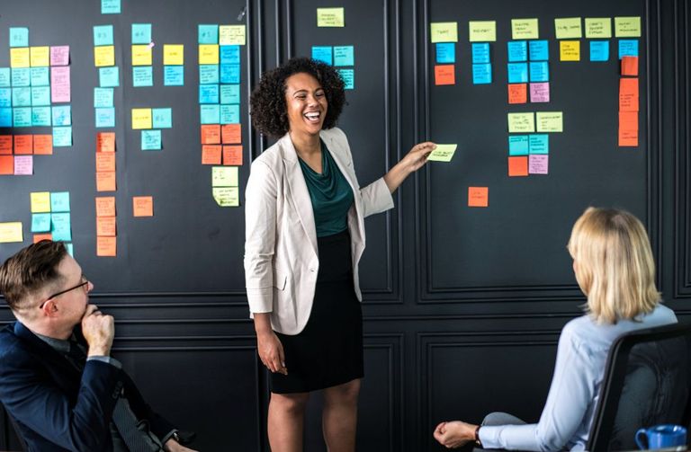 A group of three people in a collaborative meeting or brainstorming session. A woman is standing and presenting ideas using colorful sticky notes on a black wall, which is covered with a variety of notes arranged in an organized manner. She is smiling and appears to be engaging with her colleagues, who are seated and attentively listening. The atmosphere is energetic and positive, indicating a productive and creative work environment.